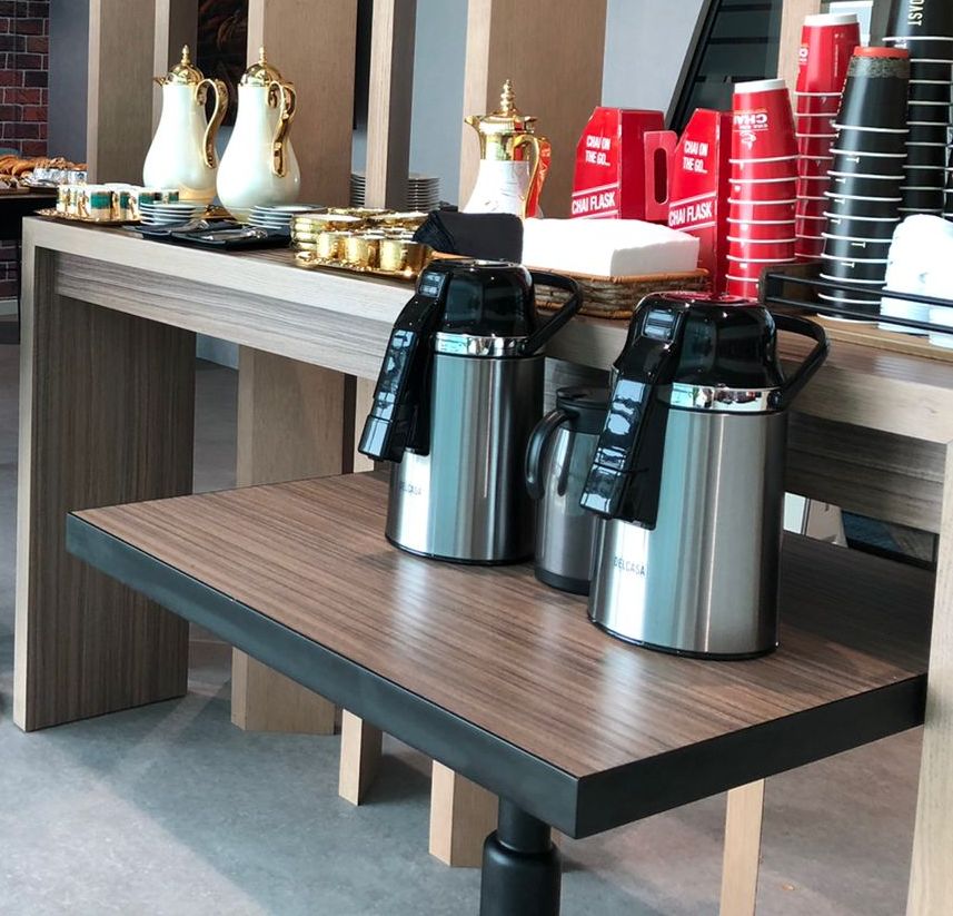 Hospitality table with two white and gold gahwa dallahs, small cups, red Chai Flask boxes, stacked red and black paper cups and two steel airpot flasks