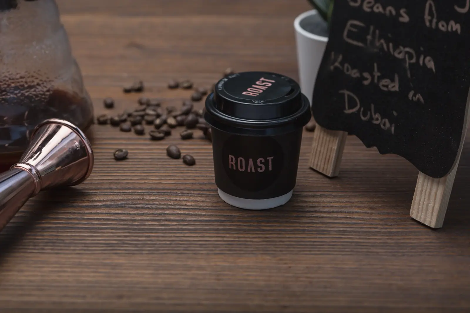ROAST takeaway cup on a wooden table with scattered coffee beans, a copper measuring cup and a small chalkboard sign